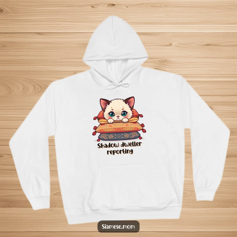 Funny Siamese cub peeking from behind soft cushions, a fluffy kitten offering comfort and humor on a warm hoodie.